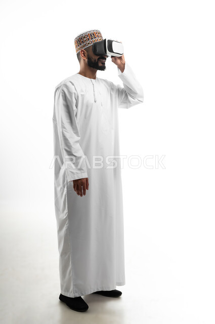 The concept of integration and simulation of the imaginary world, fun and entertainment by watching through virtual reality glasses, portrait of a young Arab Gulf Omani man wearing 3D glasses, facial gestures indicating joy and happiness, white background