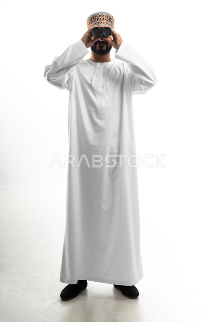 مفهوم النظرة المستقبلية، رؤية الاشياء البعيدة عبر التلسكوب, حب الاستكشاف و المغامرة, بورتريه لشاب عربي خليجي عماني مبتسم يرتدي الدشداشة و الكمه يحمل بيده منظار ثنائي العينين و يينظر الى شيئ ما, ايماءات الاستمتاع و السرور, صورة الجسم بالطول الكامل, خلفية بيضاء