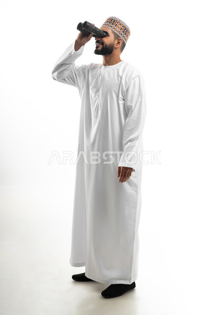 مفهوم النظرة المستقبلية، رؤية الاشياء البعيدة عبر التلسكوب, حب الاستكشاف و المغامرة, بورتريه لشاب عربي خليجي عماني مبتسم يرتدي الدشداشة و الكمه يحمل بيده منظار ثنائي العينين و يينظر الى شيئ ما, ايماءات الاستمتاع و السرور, صورة الجسم بالطول الكامل, خلفية بيضاء