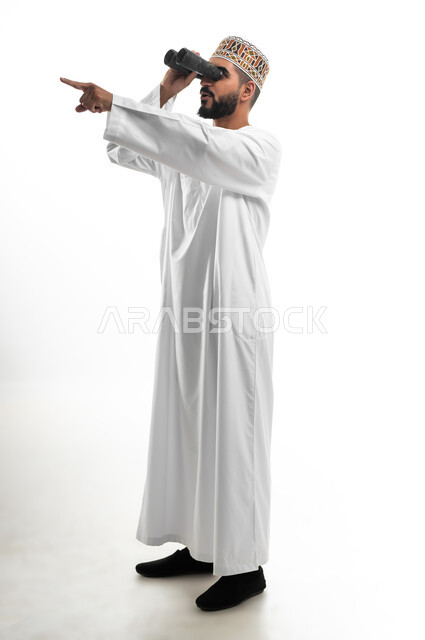 مفهوم النظرة المستقبلية، رؤية الاشياء البعيدة عبر التلسكوب, حب الاستكشاف و المغامرة, بورتريه لشاب عربي خليجي عماني مبتسم يرتدي الدشداشة و الكمه يحمل بيده منظار ثنائي العينين و يينظر الى شيئ ما, ايماءات الاستمتاع و السرور, صورة الجسم بالطول الكامل, خلفية بيضاء