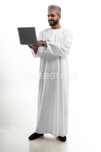 مفهوم تسهيل الاعمال والقدرة على إنجاز المهام عن بعد، استخدام التقنيات الحديثة والأجهزة المتطورة ، بورتريه لشاب عربي خليجي عماني يرتدي الدشداشة والكمه يمسك بيده حاسوب محمول تظهر عليه علامات الفرح والسرور، الجسم بالطول الكامل، خلفية بيضاء