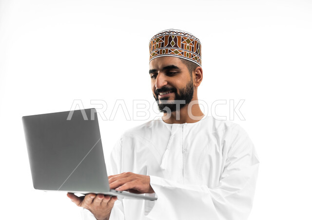 The concept of facilitating work and the ability to accomplish tasks remotely, using modern technologies and advanced devices, a portrait of a young Gulf Arab Omani man wearing a dishdasha and a sleeve, holding a laptop in his hand, showing signs of joy and happiness, white background
