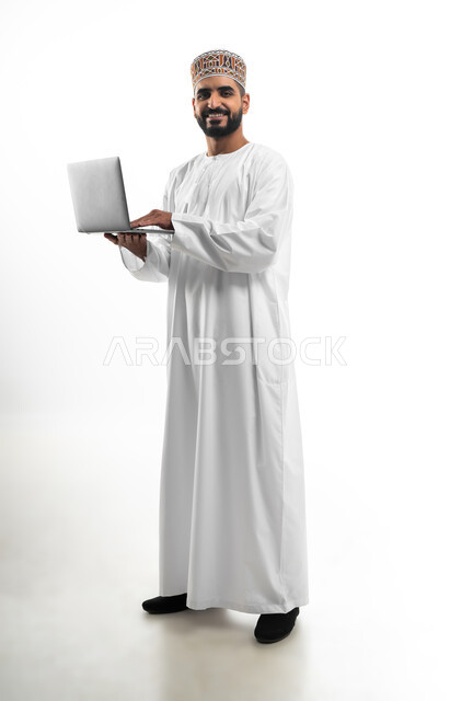 مفهوم تسهيل الاعمال والقدرة على إنجاز المهام عن بعد، استخدام التقنيات الحديثة والأجهزة المتطورة ، بورتريه لشاب عربي خليجي عماني يرتدي الدشداشة والكمه يمسك بيده حاسوب محمول تظهر عليه علامات الفرح والسرور، الجسم بالطول الكامل، خلفية بيضاء
