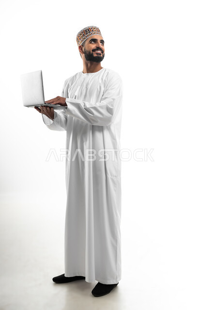مفهوم تسهيل الاعمال والقدرة على إنجاز المهام عن بعد، استخدام التقنيات الحديثة والأجهزة المتطورة ، بورتريه لشاب عربي خليجي عماني يرتدي الدشداشة والكمه يمسك بيده حاسوب محمول تظهر عليه علامات الفرح والسرور، الجسم بالطول الكامل، خلفية بيضاء