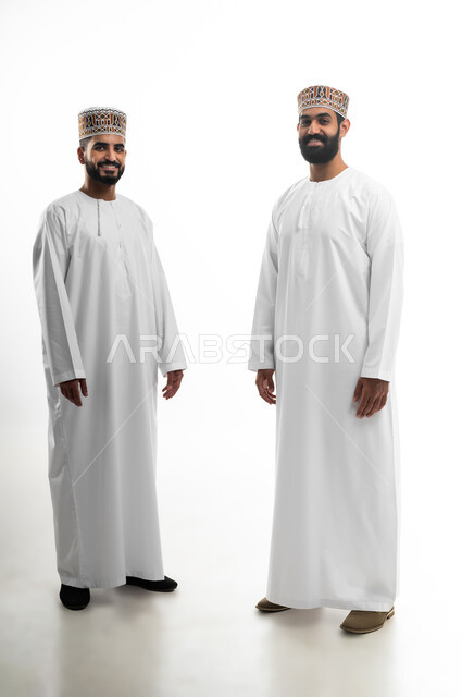 مفهوم الاهتمام بالمظهر الخارجي والأناقة والنظافة الشخصية، النظر للكاميرا بتعابير السعادة والسرور، بورتريه لشابان عربيان خليجيان عمانيان يرتديان الدشداشة والكمه تظهر عليهما علامات الفرح، الجسم بالطول الكامل، خلفية بيضاء