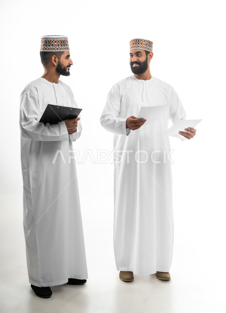 النقاش والحوار حول موضوع ما، تدوين الملاحظات ومراجعة المعلومات، بورتريه لشابان عربيان خليجيان عمانيان يرتديان الدشداشة والكمه تظهر عليهما تعابير الفرح والسرور، مفهوم الجد والاجتهاد في العمل، خلفية بيضاء