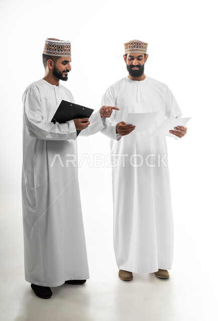 Discussion and dialogue on a topic, taking notes and reviewing information, portrait of two young Gulf Arab Omani men wearing dishdasha and kummah, expressions of joy and happiness appear on them, concept of seriousness and diligence in work, white background