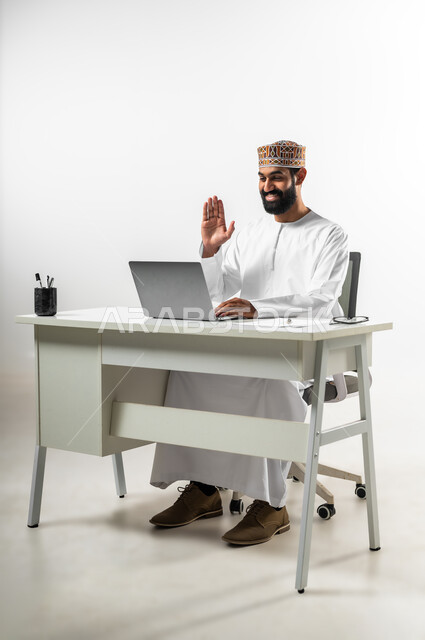 إجراء مكالمة فيديو عبر الحاسوب المحمول، استخدام التقنيات الحديثة في التواصل مع العملاء، بورتريه لرجل عربي عماني يرتدي الدشداشة والكمه يجلس في مكتبه يجري محادثة عبر الانترنت، خلفية بيضاء