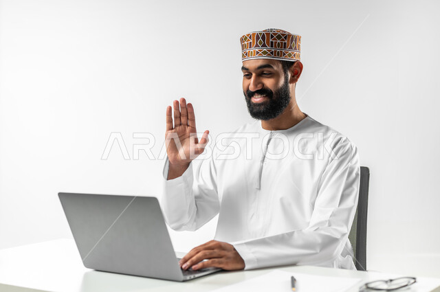 Making a video call via laptop, using modern technologies in communicating with clients, portrait of an Omani Arab man wearing a dishdasha and a turban sitting in his office having a conversation over the internet, white background