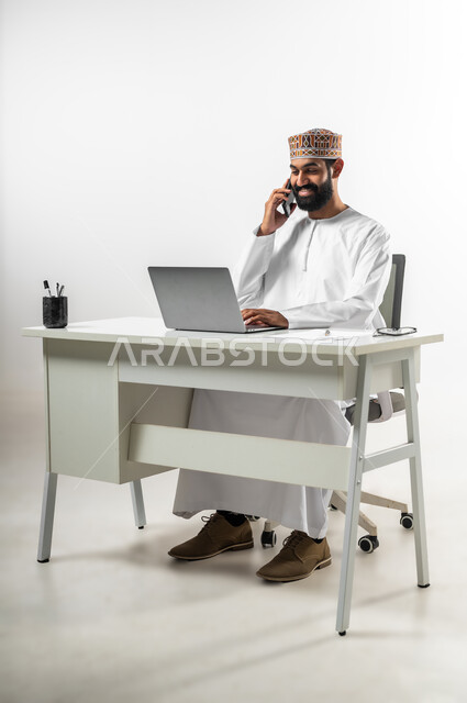 استعمال الهاتف المحمول لإجراء مكالمات هاتفية مع العملاء، بورتريه لشاب عربي خليجي عماني يرتدي الدشداشة والكمه يجلس خلف طاولة مكتبه تظهر عليه علامات الفرح والسرور، استخدام الأجهزة الحديثة والتقنيات المتطورة، خلفية بيضاء