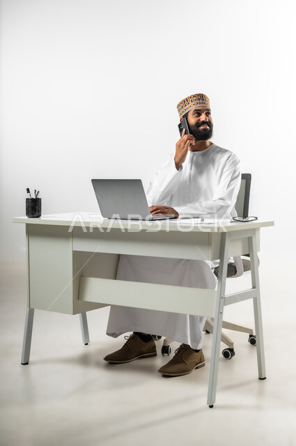 Using a mobile phone to make phone calls to clients, portrait of a young Omani Gulf Arab man wearing a dishdasha and a kummah sitting behind his office desk, showing signs of joy and happiness, using modern devices and advanced technologies, white background