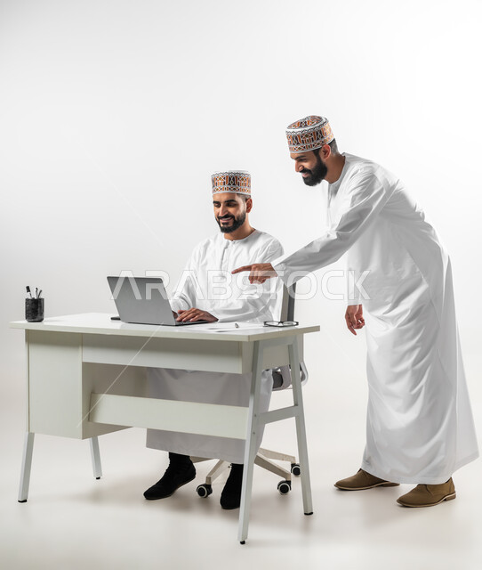 المساعدة والمشاركة في إنجاز الأعمال، اللطف والرقي في التعامل بين الموظفين، بورتريه لشابان عربيان خليجيان عمانيان يرتديان الدشداشة والكمه يقومان بإنهاء العمل سوياً، تعابير وجه تدل على الفرح والسرور، خلفية بيضاء