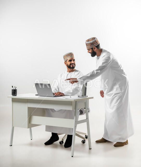 Help and participation in completing work, kindness and sophistication in dealing between employees, portrait of two young Gulf Arab Omani men wearing a dishdasha and a kummah completing work together, facial expressions indicating joy and happiness, white background