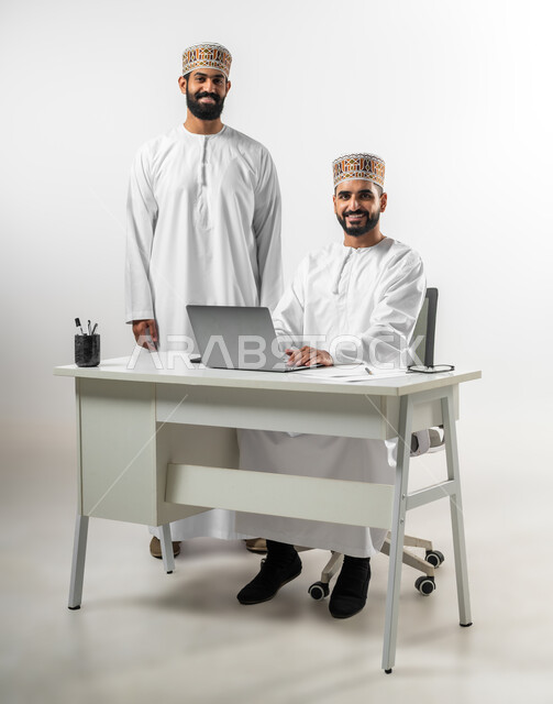 النظر للكاميرا بتعابير السعادة والسرور، الاستعداد لبدء العمل وإنجاز المهام المطلوبة، بورتريه لشابان عربيان خليجيان عمانيان يرتديان الدشداشة والكمه أحدهما جالس والآخر يقف ينظران بابتسامة لطيفة، خلفية بيضاء