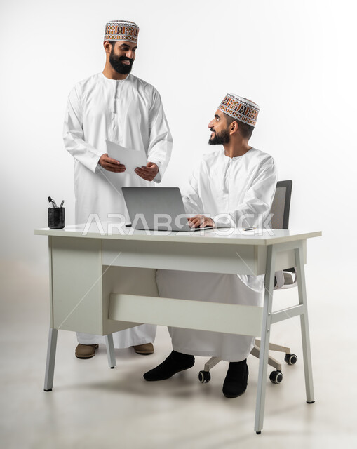 إعطاء التعليمات وشرح المهام، الرقي واللطف في التعامل بين الموظفين، بورتريه لشابان عربيان خليجيان عمانيان يرتديان الدشداشة والكمه يقومان بالمشاركة في العمل، تعابير وجه تدل على الفرح والسرور، خلفية بيضاء