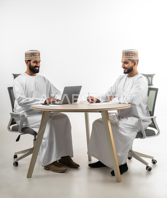 الدردشة والمشاركة في إنجاز المهام المطلوبة، الصحبة وتبادل الأحاديث والخبرات، بورتريه لشابان عربيان خليجيان عمانيان يرتديان الدشداشة والكمه تظهر عليهما علامات الفرح والسرور، استخدم التقنيات الحديثة لإنهاء الأعمال، خلفية بيضاء