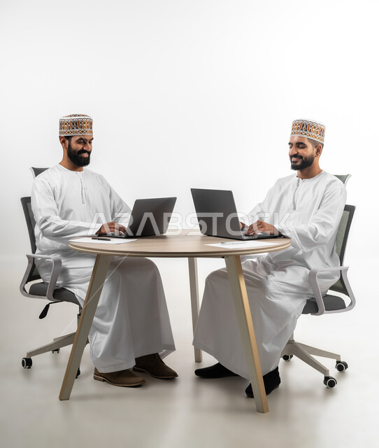 تعابير الفرح والسرور، استخدام الأجهزة المتطورة والتقنيات الحديثة لإنجاز الأعمال، بورتريه لشابان عربيان خليجيان عمانيان يرتديان الدشداشة والكمه يتشاركان في طاولة العمل  تظهر عليهما علامات السرور، خلفية بيضاء