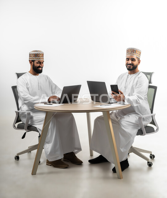 تعابير الفرح والسرور، استخدام الأجهزة المتطورة والتقنيات الحديثة لإنجاز الأعمال، بورتريه لشابان عربيان خليجيان عمانيان يرتديان الدشداشة والكمه يتشاركان في طاولة العمل  تظهر عليهما علامات السرور، خلفية بيضاء