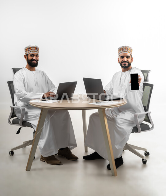 تعابير الفرح والسرور، استخدام الأجهزة المتطورة والتقنيات الحديثة لإنجاز الأعمال، بورتريه لشابان عربيان خليجيان عمانيان يرتديان الدشداشة والكمه يتشاركان في طاولة العمل  تظهر عليهما علامات السرور، خلفية بيضاء