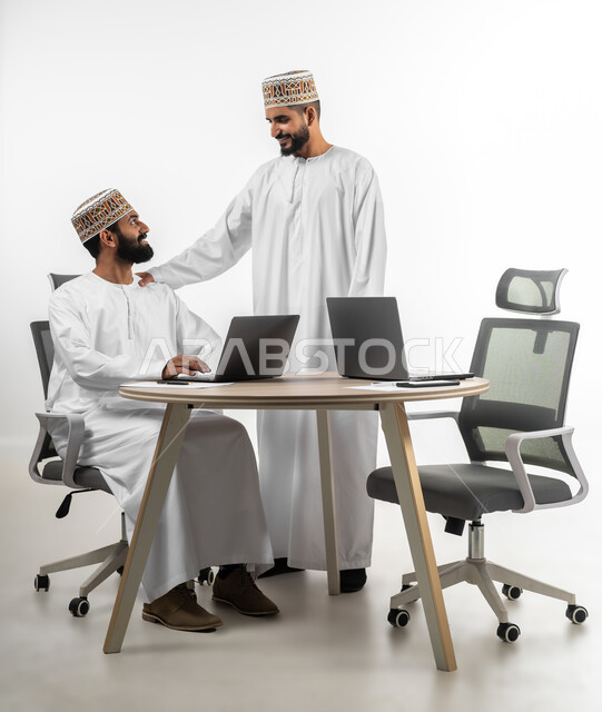 المساعدة والمشاركة في إنجاز الأعمال، اللطف والرقي في التعامل بين الموظفين، بورتريه لشابان عربيان خليجيان عمانيان يرتديان الدشداشة والكمه يقومان بإنهاء العمل سوياً، تعابير وجه تدل على الفرح والسرور، خلفية بيضاء