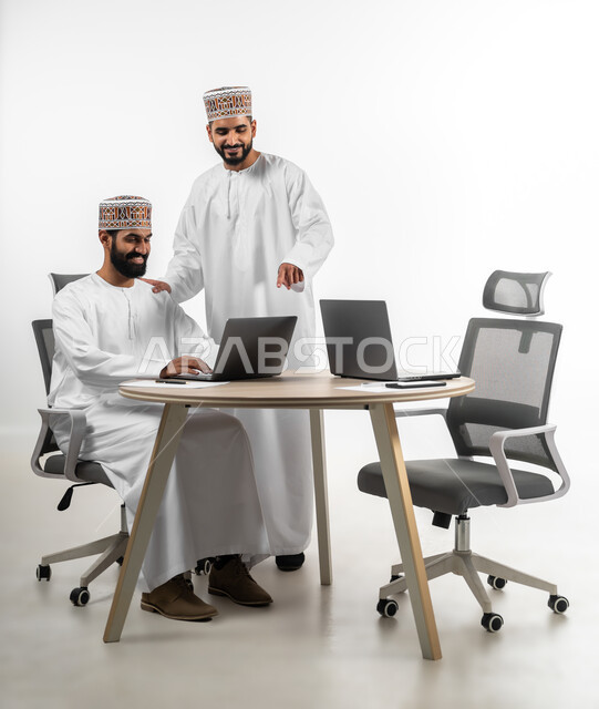 المساعدة والمشاركة في إنجاز الأعمال، اللطف والرقي في التعامل بين الموظفين، بورتريه لشابان عربيان خليجيان عمانيان يرتديان الدشداشة والكمه يقومان بإنهاء العمل سوياً، تعابير وجه تدل على الفرح والسرور، خلفية بيضاء