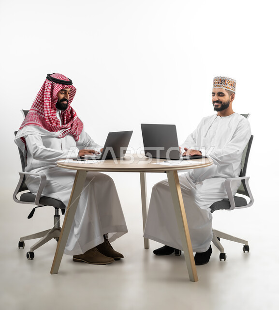 الجلوس على طاولة عمل مشتركة وإنجاز المهام، الحوار والمناقشة وتبادل المعلومات، بورتريه لشابان عربيان سعودي وعماني يجلسان مقابل بعضهما بايماءات السرور والفرح، الإحساس بالمتعة والسعادة أثناء القيام بالأعمال المطلوبة، خلفية بيضاء