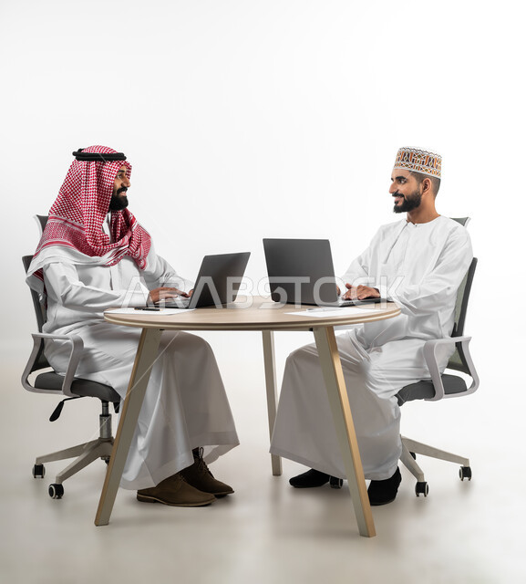 Sitting at a shared work table and completing tasks, dialogue, discussion and exchange of information, portrait of two young Arab men, Saudi and Omani, sitting opposite each other with gestures of pleasure and joy, feeling pleasure and happiness while doing the required work, white background