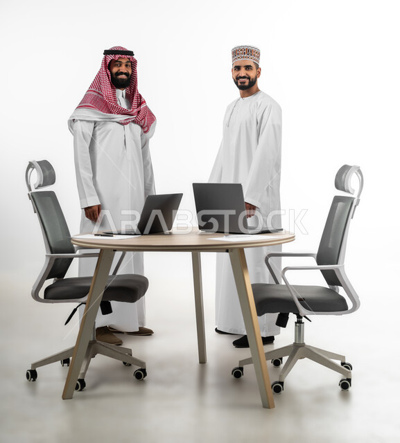 النظر للكاميرا بتعابير السعادة والسرور، الاستعداد لبدء العمل وإنجاز المهام المطلوبة، بورتريه لرجل عربي خليجي سعودي وآخر عماني يقفان مقابل بعضهما ينظران بابتسامة لطيفة، الجسم بالطول الكامل، خلفية بيضاء