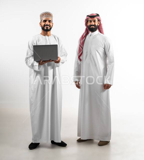 المشاركة في إنجاز الأعمال، مفهوم العمل والاجتهاد، بورتريه لشاب عماني يرتدي الدشداشة والكمه يحمل بيده حاسب محمول وآخر سعودي تظهر عليهما علامات الفرح والسرور، الجسم بالطول الكامل، تبادل الثقافات بين الدول العربية، خلفية بيضاء