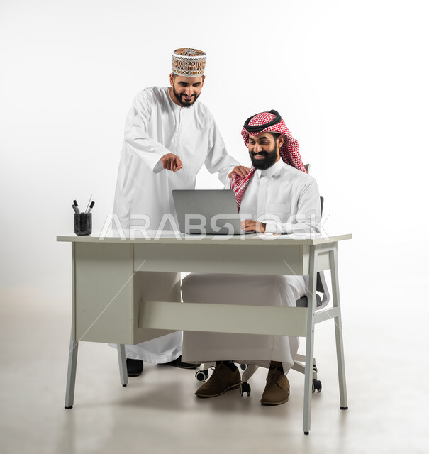 المساعدة في إنجاز الاعمال، بورتريه لشابان عربيان سعودي وعماني يرتدي كل منهما الزي التقليدي الخاص ببلده يتشاركان في إنهاء المهام، استخدام التقنيات الحديثة في مجال العمل، ايماءات السعادة والسرور، خلفية بيضاء