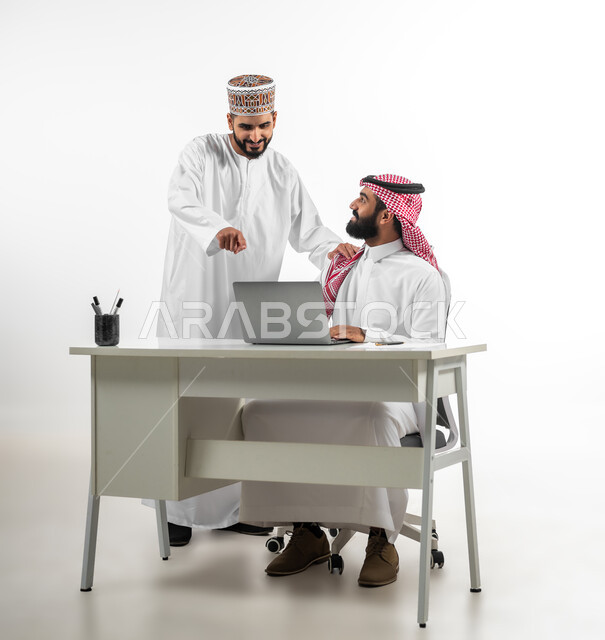 المساعدة في إنجاز الاعمال، بورتريه لشابان عربيان سعودي وعماني يرتدي كل منهما الزي التقليدي الخاص ببلده يتشاركان في إنهاء المهام، استخدام التقنيات الحديثة في مجال العمل، ايماءات السعادة والسرور، خلفية بيضاء