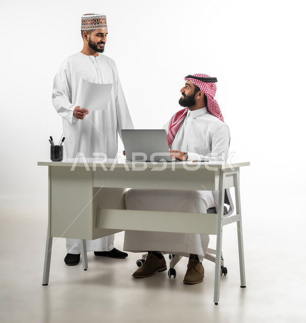 Work atmosphere and accomplishing tasks, portrait of an Omani Arab young man wearing a dishdasha and a kuma explaining the nature of the business to a Saudi man wearing a shemakh, kindness and sophistication in dealing with employees, gestures of happiness and pleasure, white background