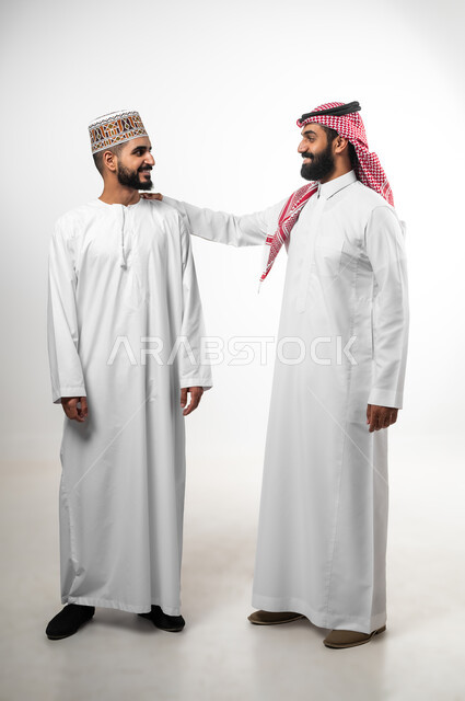 الدردشة مع الأصدقاء، الرقي في التعامل وتبادل الأحاديث، بورتريه لرجل عربي سعودي يرتدي الدشداشة والشماخ يضع يده على كتف شاب عماني تظهر عليهما علامات الفرح والسرور، الجسم بالطول الكامل، خلفية بيضاء