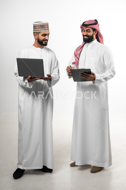 المشاركة في إنجاز الأعمال، مفهوم العمل والاجتهاد، بورتريه لشاب عماني يرتدي الدشداشة والكمه وآخر سعودي تظهر عليهما علامات الفرح والسرور، الجسم بالطول الكامل، تبادل الثقافات بين الدول العربية، خلفية بيضاء
