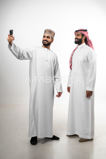 استخدام الهاتف المحمول لالتقاط صورة سيلفي، بورتريه لشابان عربيان سعودي وعماني يرتدي كل منهما الزي الخاص ببلده يقومان بتوثيق اللحظات، النظر للكاميرا بتعابير السعادة والسرور، الطول الكامل للجسم، خلفية بيضاء