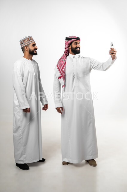 استخدام الهاتف المحمول لالتقاط صورة سيلفي، بورتريه لشابان عربيان سعودي وعماني يرتدي كل منهما الزي الخاص ببلده يقومان بتوثيق اللحظات، النظر للكاميرا بتعابير السعادة والسرور، الطول الكامل للجسم، خلفية بيضاء