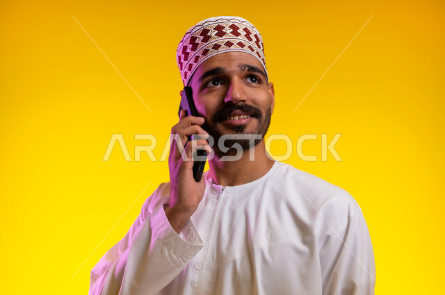 الدردشة مع الأهل والأصدقاء، تسهيل التواصل مع الآخرين باستخدام الجوال، بورتريه لرجل عربي خليجي عماني يرتدي الدشداشة والكمه يقوم بالتحدث مع الأهل والاصدقاء باستخدام الهاتف المحمول، خلفيه برتقاليه