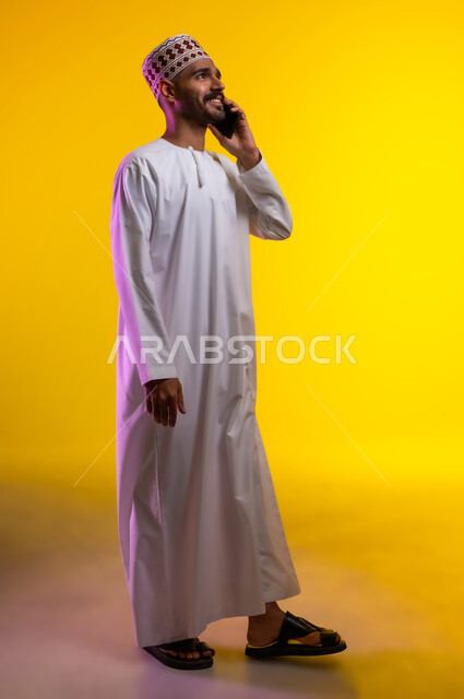 الدردشة مع الأهل والأصدقاء، تسهيل التواصل مع الآخرين باستخدام الجوال، بورتريه لرجل عربي خليجي عماني يرتدي الدشداشة والكمه يقوم بالتحدث مع الأهل والاصدقاء باستخدام الهاتف المحمول، خلفيه برتقاليه