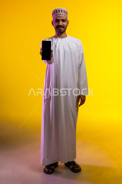 التقاط الصور التذكارية وتوثيق اللحظات السعيدة, بورتريه لرجل عربي خليجي عماني يرتدي الدشداشه والكمه يمسك بيده الهاتف المحمول ويشير بشاشه سوداء فارغه، مفهوم تطبيقات الجوال الاون لاين والتواصل الاجتماعي عن بعد، خلفية برتقاليه