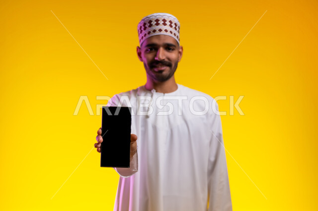 التقاط الصور التذكارية وتوثيق اللحظات السعيدة, بورتريه لرجل عربي خليجي عماني يرتدي الدشداشه والكمه يمسك بيده الهاتف المحمول ويشير بشاشه سوداء فارغه، مفهوم تطبيقات الجوال الاون لاين والتواصل الاجتماعي عن بعد، خلفية برتقاليه