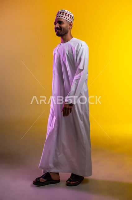 مفهوم الاناقة و الاهتمام بالمظهر الخارجي, بورتريه  لشاب عربي خليجي عماني يرتدي الدشداشة و الكمه يقف وينظر بإيماءات السعادة و السرور, تعابير السير و المشي للذهاب الى مكان ما, الوقوف والنظر الى الامام، صورة الجسم بالطول الكامل, خلفيه برتقاليه