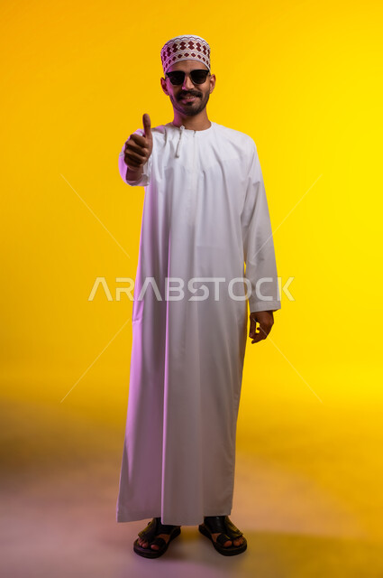 رفع إصبع الإبهام للأعلى دليل على الإعجاب، ايماءات تدل على الموافقة والقبول، بورتريه لرجل عربي خليجي عماني يرتدي الدشداشة والكمه تظهر عليه تعابير الفرح والسرور، الرضى وعدم الرفض، خلفية برتقاليه