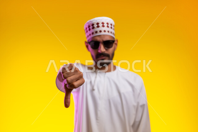 مفهوم الرفض وعدم القبول، بورتريه لشاب عربي خليجي عماني يرتدي الدشداشة و الكمه يشير بإصبع الابهام للاسفل بإيماءات عدم الاعجاب والتقييم السلبي، الشعور بالانزعاج من شيء ما، خلفية برتقاليه