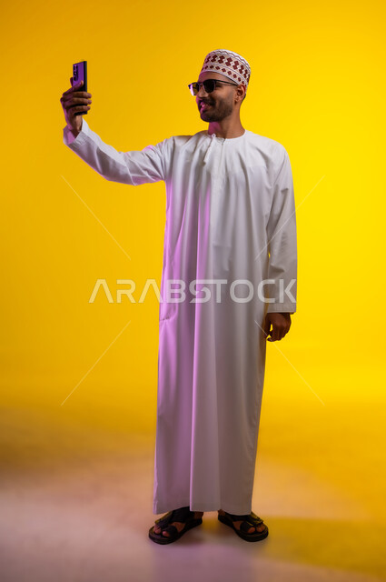 التقاط الصور التذكارية وتوثيق اللحظات السعيدة, بورتريه لرجل عربي خليجي عماني يرتدي الدشداشه والكمه ويضع النظارات الشمسيه يمسك الهاتف المحمول ويقوم بألتقاط صورة سيلفي لنفسه, مفهوم الأناقة والرجولة والاهتمام بالمظهر الخارجي, خلفية برتقاليه