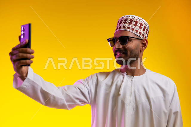 Taking souvenir photos and documenting happy moments, a portrait of an Arab Gulf Omani man wearing a dishdasha, a sleeve, and wearing sunglasses, holding a mobile phone and taking a selfie of himself, the concept of elegance, masculinity, and interest in external appearance, orange background