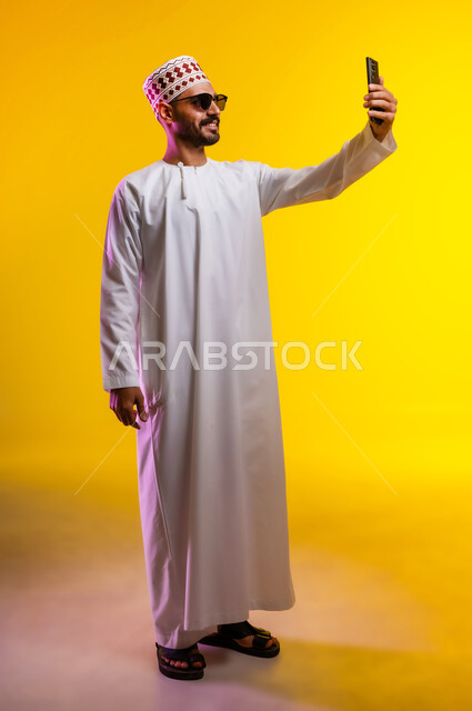 التقاط الصور التذكارية وتوثيق اللحظات السعيدة, بورتريه لرجل عربي خليجي عماني يرتدي الدشداشه والكمه ويضع النظارات الشمسيه يمسك الهاتف المحمول ويقوم بألتقاط صورة سيلفي لنفسه, مفهوم الأناقة والرجولة والاهتمام بالمظهر الخارجي, خلفية برتقاليه