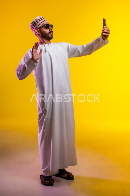 التقاط الصور التذكارية وتوثيق اللحظات السعيدة, بورتريه لرجل عربي خليجي عماني يرتدي الدشداشه والكمه ويضع النظارات الشمسيه يمسك الهاتف المحمول ويقوم بألتقاط صورة سيلفي لنفسه, مفهوم الأناقة والرجولة والاهتمام بالمظهر الخارجي, خلفية برتقاليه