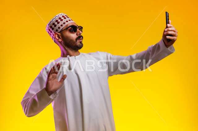 Taking souvenir photos and documenting happy moments, a portrait of an Arab Gulf Omani man wearing a dishdasha, a sleeve, and wearing sunglasses, holding a mobile phone and taking a selfie of himself, the concept of elegance, masculinity, and interest in external appearance, orange background