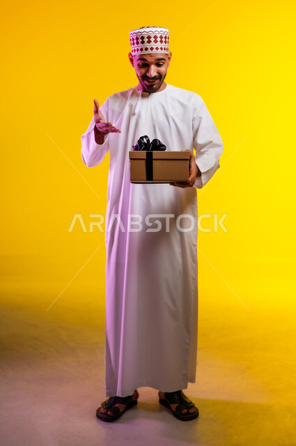ايماءات وجه تدل على الحماس لمعرفه ما بداخل صندوق الهدايا، بورتريه لرجل عربي خليجي عماني يرتدي الدشداشة والكمه يمسك بيده هديه كرتونيه يقوم بمحاوله فتحها، تعابير فرح وسرور واكتشاف المفاجات السعيده، هدايا عيد الاب ومناسبات سعيده، خلفيه برتقاليه