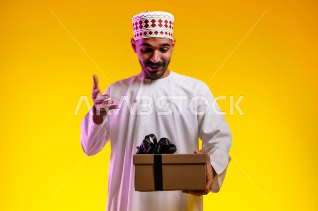 Facial gestures indicating enthusiasm to know what is inside the gift box, a portrait of an Arab Gulf Omani man wearing a dishdasha and a cuff holding a cartoon gift in his hand that he is trying to open, expressions of joy, pleasure and discovery of happy surprises, Father’s Day gifts and happy occasions, orange background